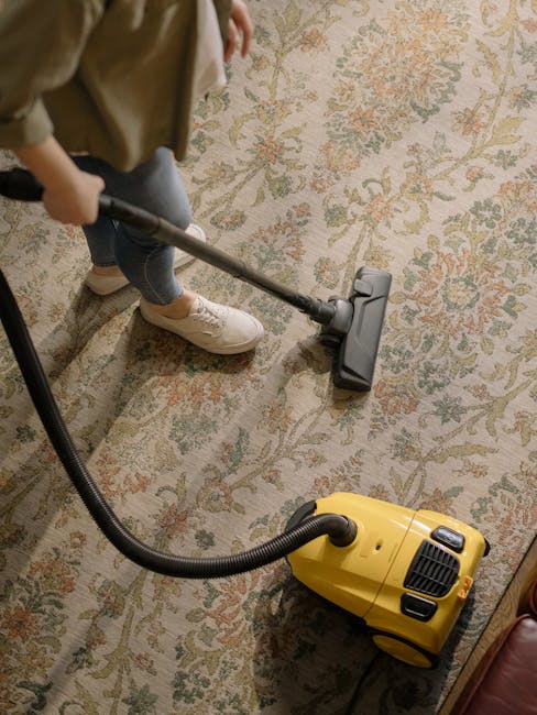 A person wearing beige shoes and blue jeans is operating a vacuum cleaner with a long black hose attachment, cleaning a patterned area rug with floral motifs in light beige, green, and muted orange to
