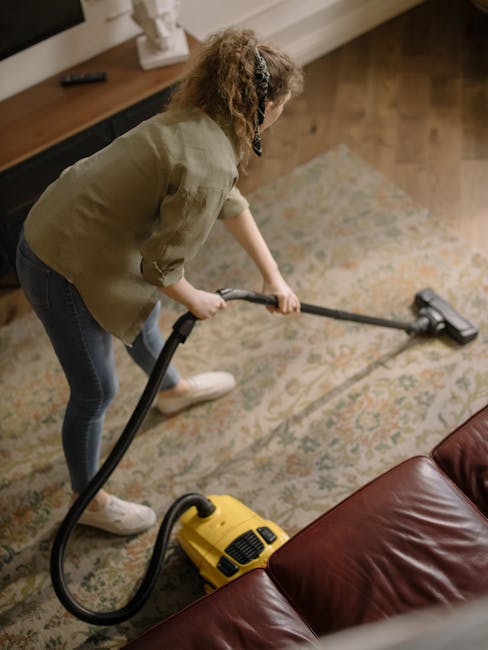 A person wearing beige shoes and blue jeans is operating a vacuum cleaner with a long black hose attachment, cleaning a patterned area rug with floral motifs in light beige, green, and muted orange to
