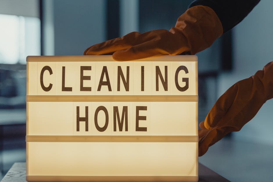 A residential living room corner features a round black side table with a rose gold trim, on which are bright yellow rubber cleaning gloves, a pink microfiber cloth, and a clear spray bottle with a gr